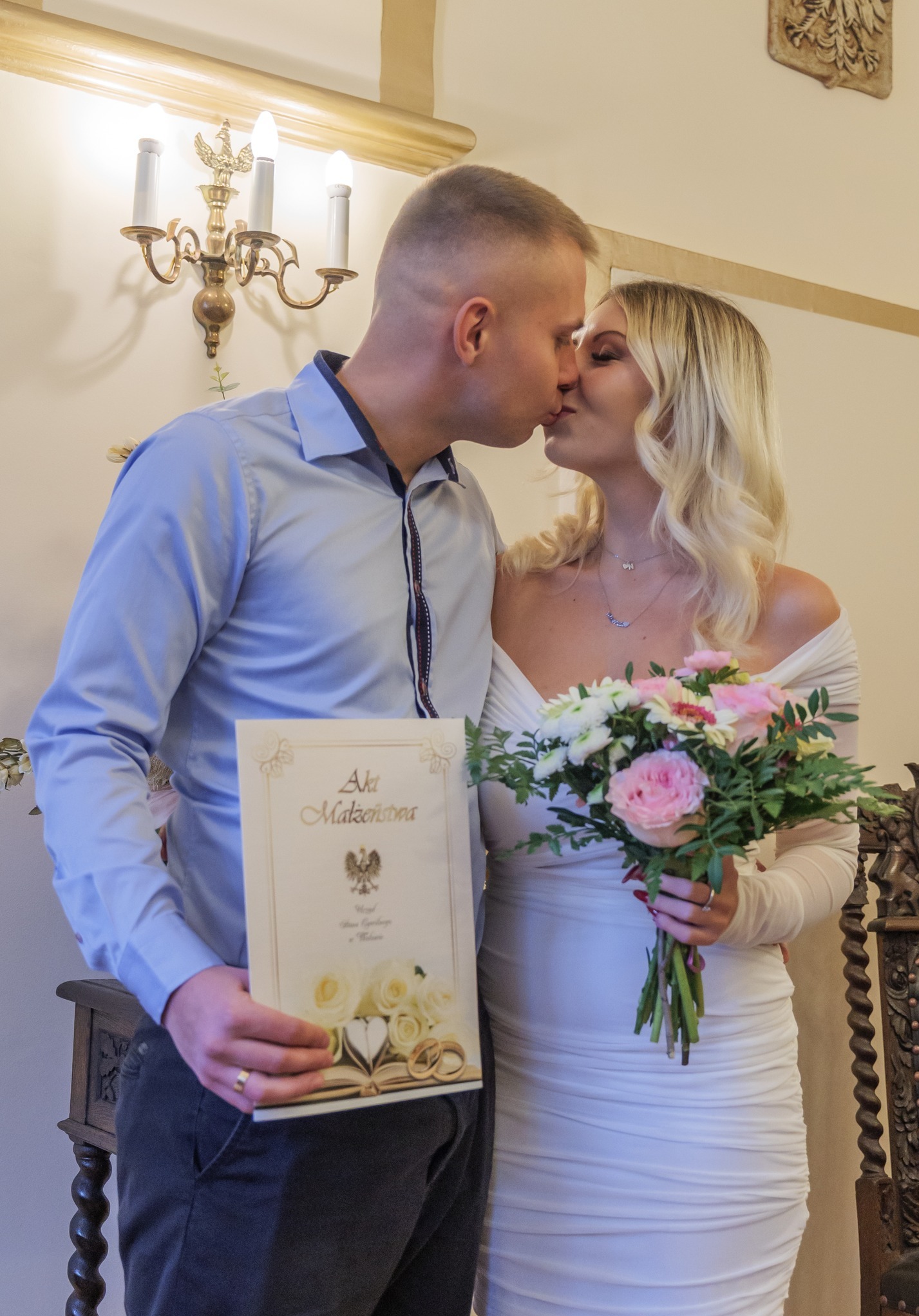 Pocałunek pary młodej trzymającej akt małżeństwa i bukiet w jasnym wnętrzu z ozdobnym żyrandolem.