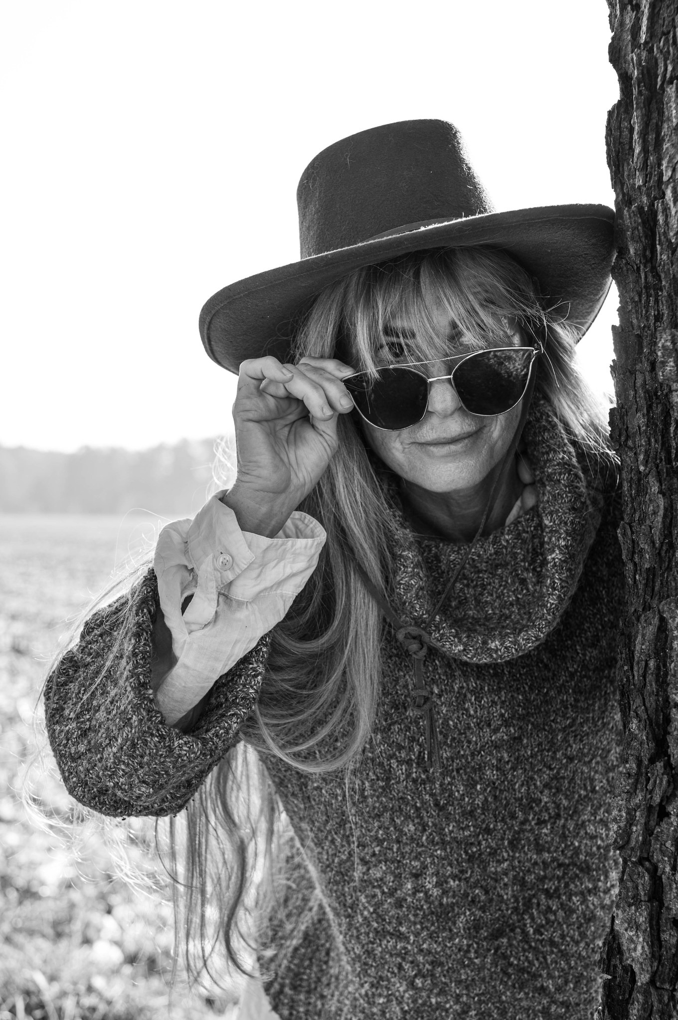 Czarno-białe zdjęcie kobiety w kapeluszu i okularach przeciwsłonecznych, opierającej się o drzewo, z rozpiętą koszulą pod swetrem