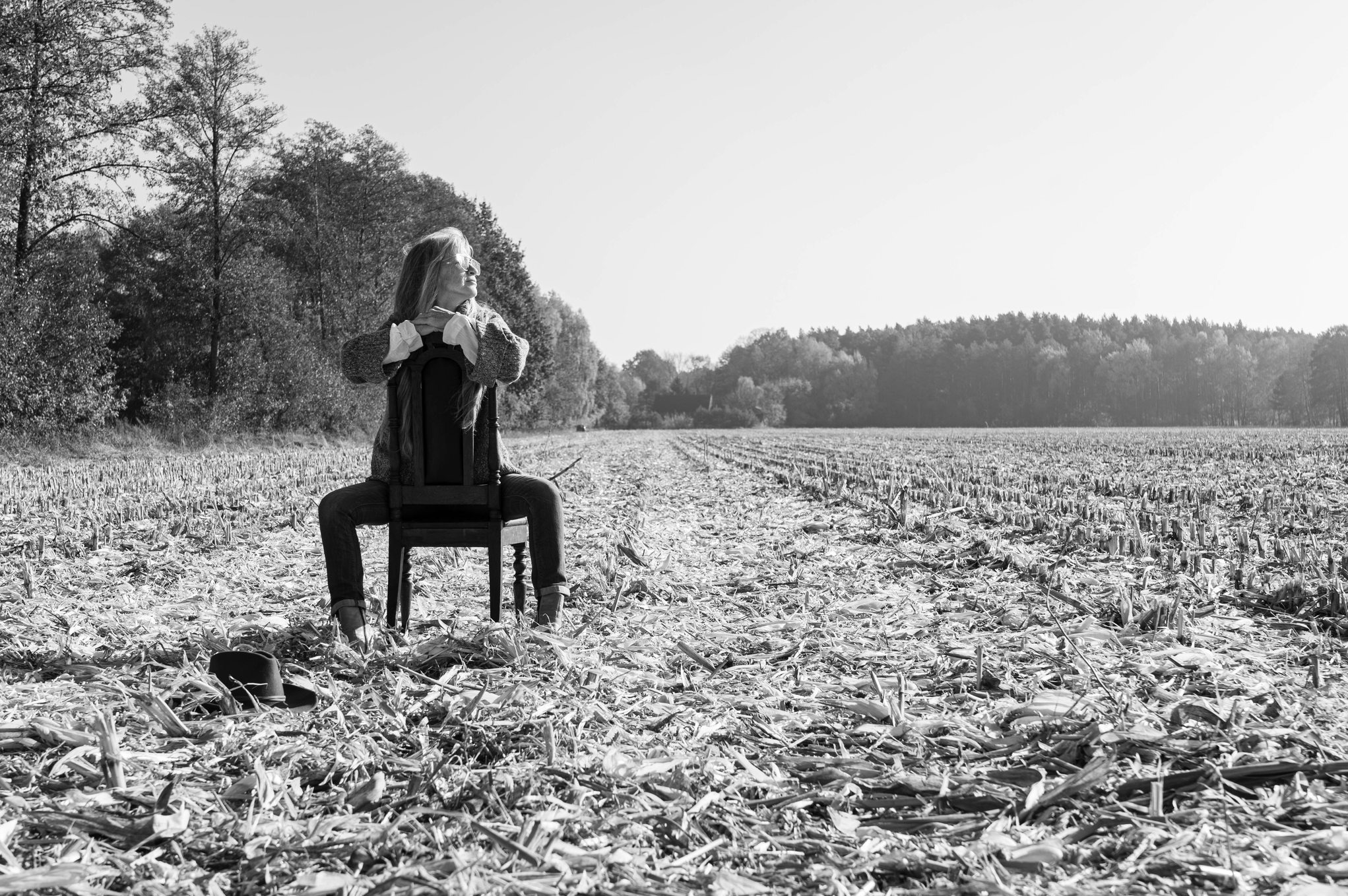 Czarno-białe zdjęcie kobiety siedzącej na krześle pośrodku zaoranego pola, w tle las, obok krzesła leży kapelusz.