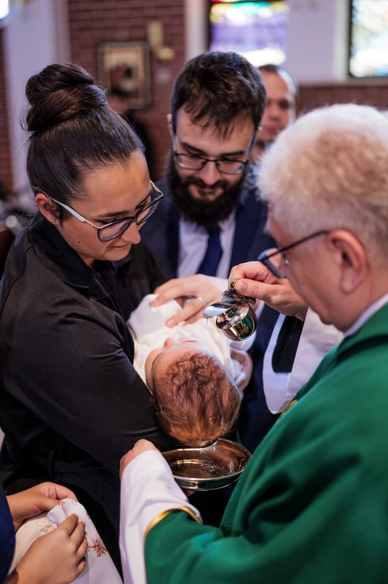 Ksiądz polewa wodą głowę niemowlęcia podczas ceremonii chrztu, rodzice dziecka obserwują z troską.