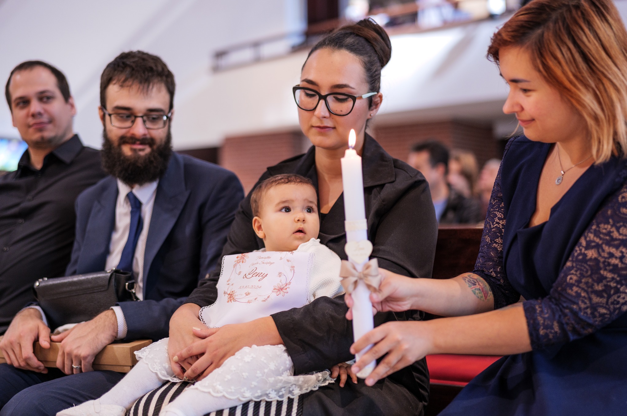 Rodzina podczas uroczystości chrztu: matka trzyma dziecko w białej sukience i z haftowanym śliniakiem, obok płonąca świeca, w tle ojciec i inni goście.