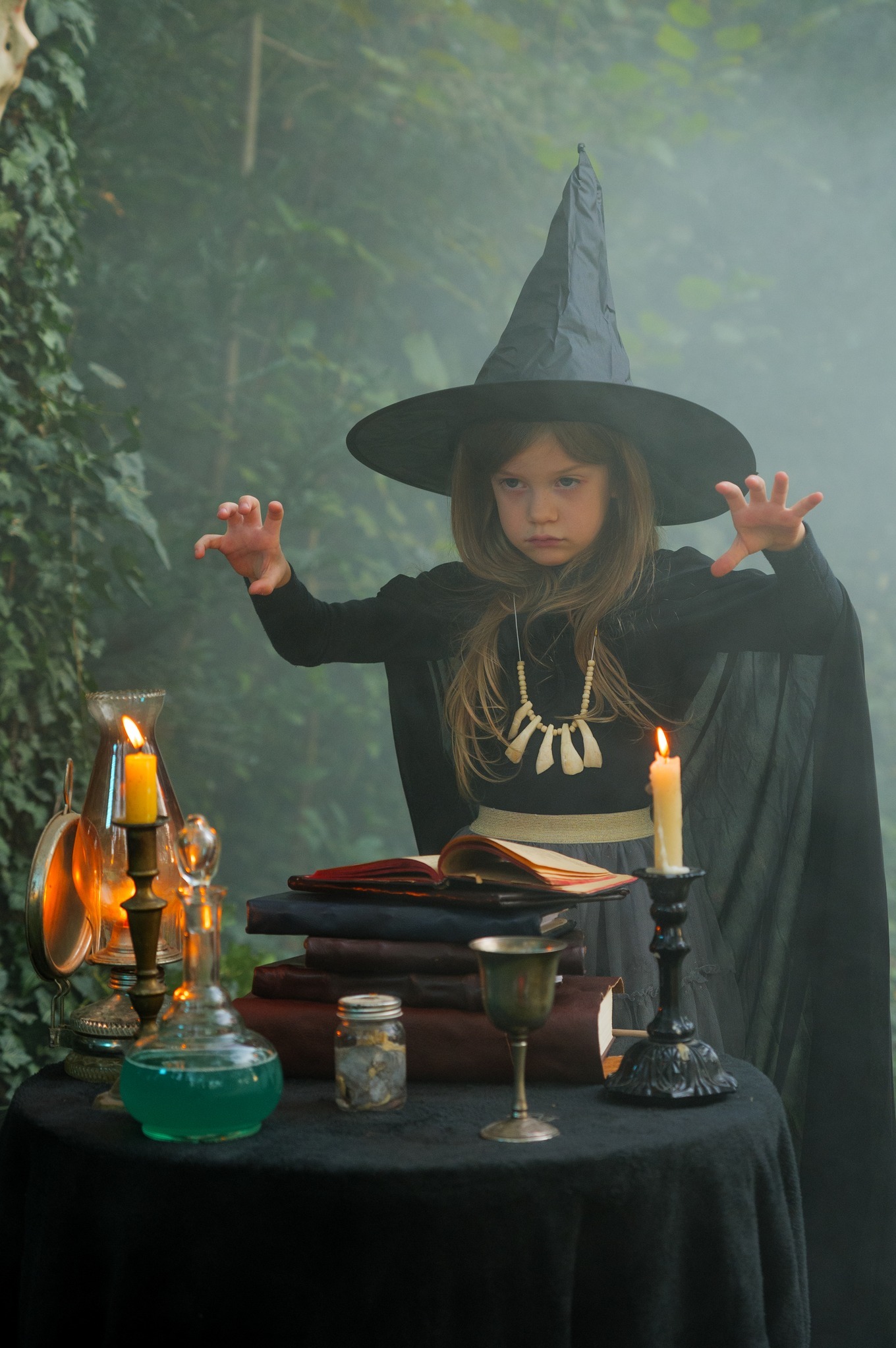 Dziewczynka w kostiumie czarownicy, z kapeluszem i naszyjnikiem z kości, stoi przy stole z eliksirami i świecami, w tle zarośnięty ogród.