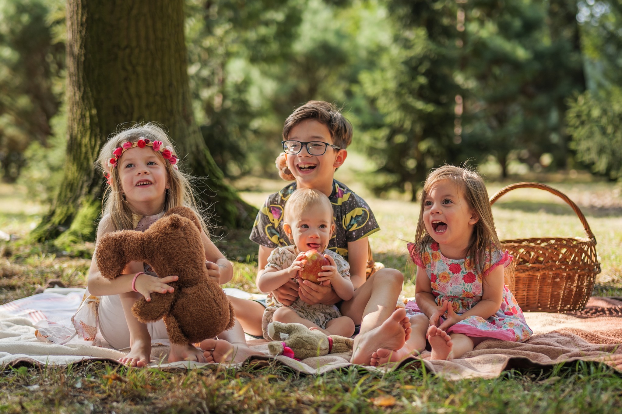 Czterech roześmianych dzieci siedzi na kocu piknikowym w lesie, jedno trzyma jabłko, drugie pluszowego misia, a w tle widoczny jest wiklinowy koszyk.