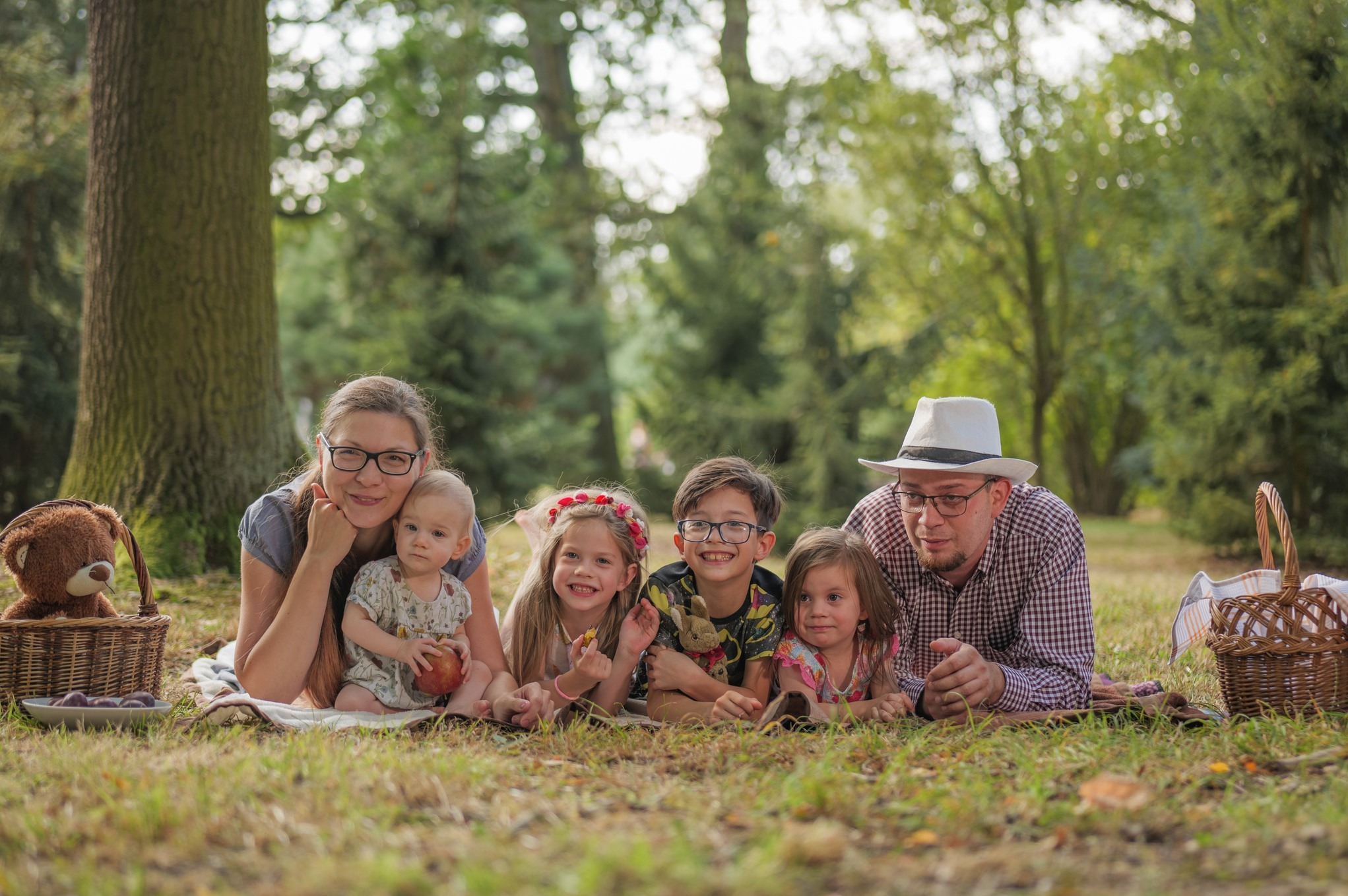 Rodzinna sesja zdjęciowa we Wrocławiu: Mama z dzieckiem trzymającym jabłko, tata w kapeluszu oraz troje uśmiechniętych dzieci leżą na kocu w parku, obok kosz piknikowy z pluszowym misiem.