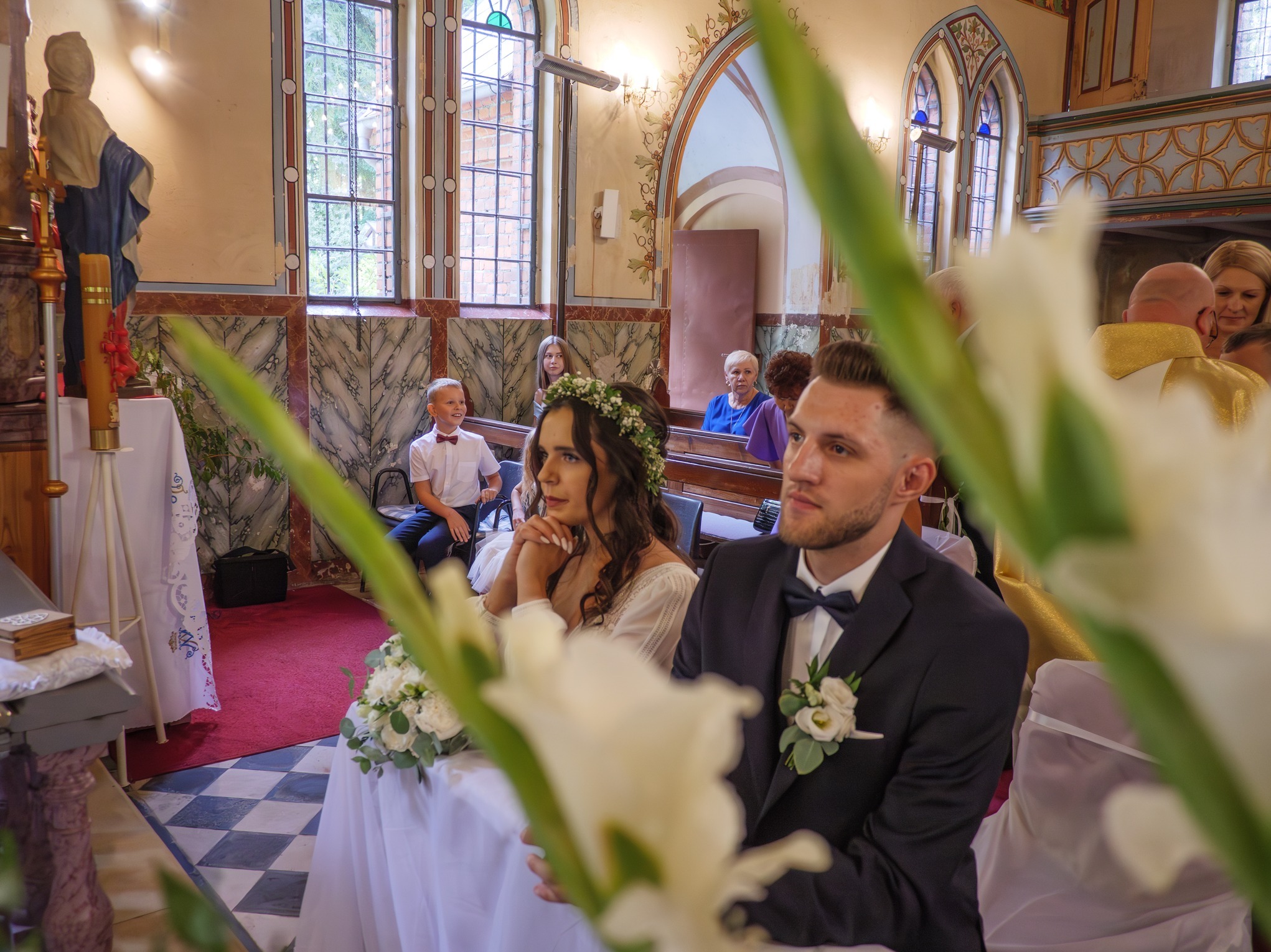 Para młoda w kościele podczas ceremonii ślubnej, widok zza kwiatów, z gośćmi w tle.