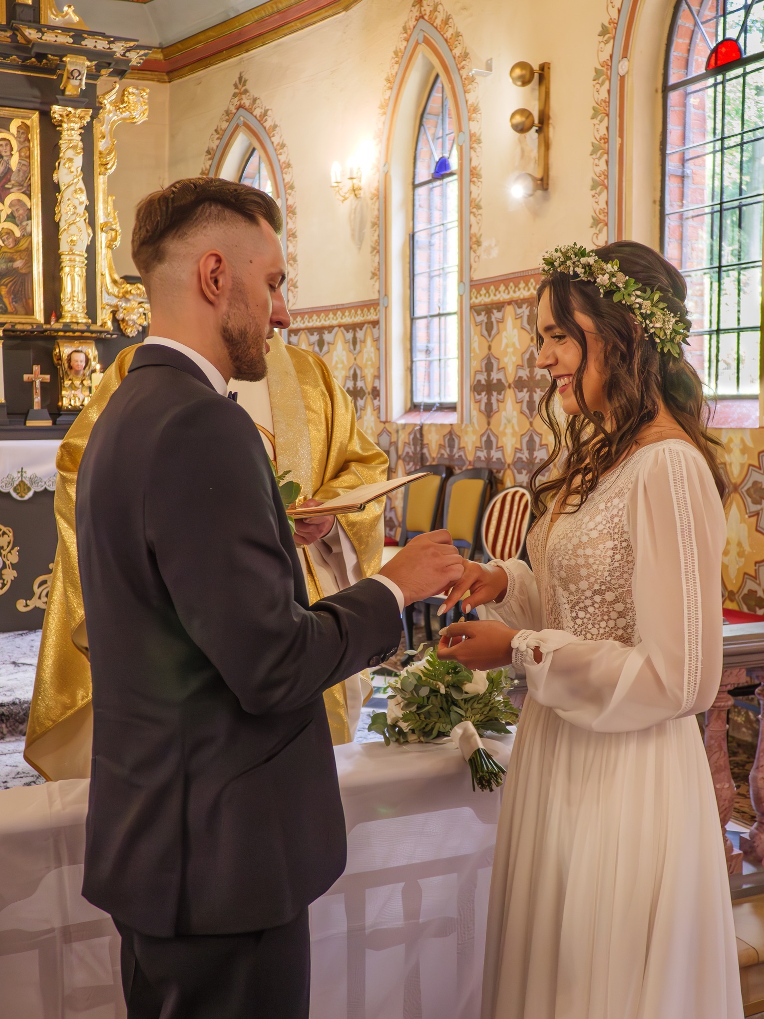 Wymiana obrączek podczas ceremonii ślubnej w kościele: panna młoda w białej sukni z koronkową górą i wiankiem, pan młody w ciemnym garniturze, ksiądz w złotej szacie, bukiet białych kwiatów na stole.