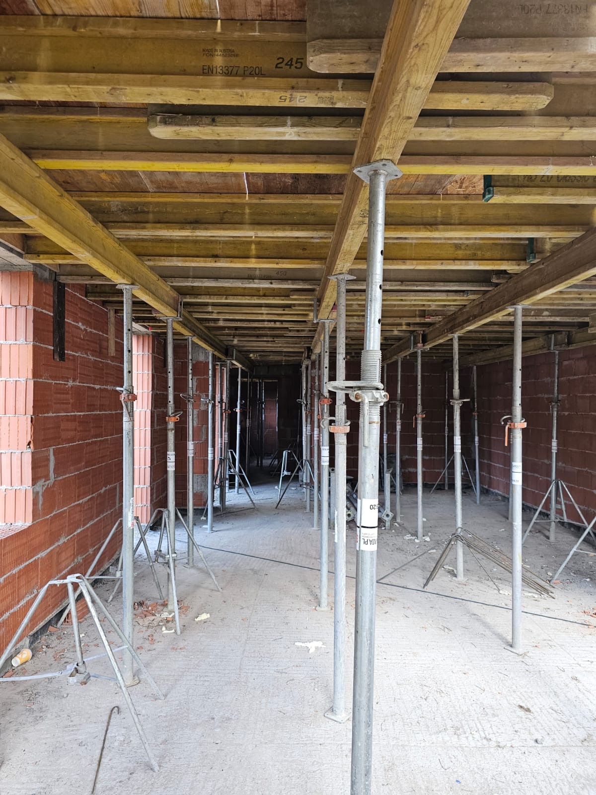 Wnętrze w trakcie budowy, widoczne czerwone cegły ścian, drewniane deski stropu podparte metalowymi stemplami, perspektywa w głąb budynku.