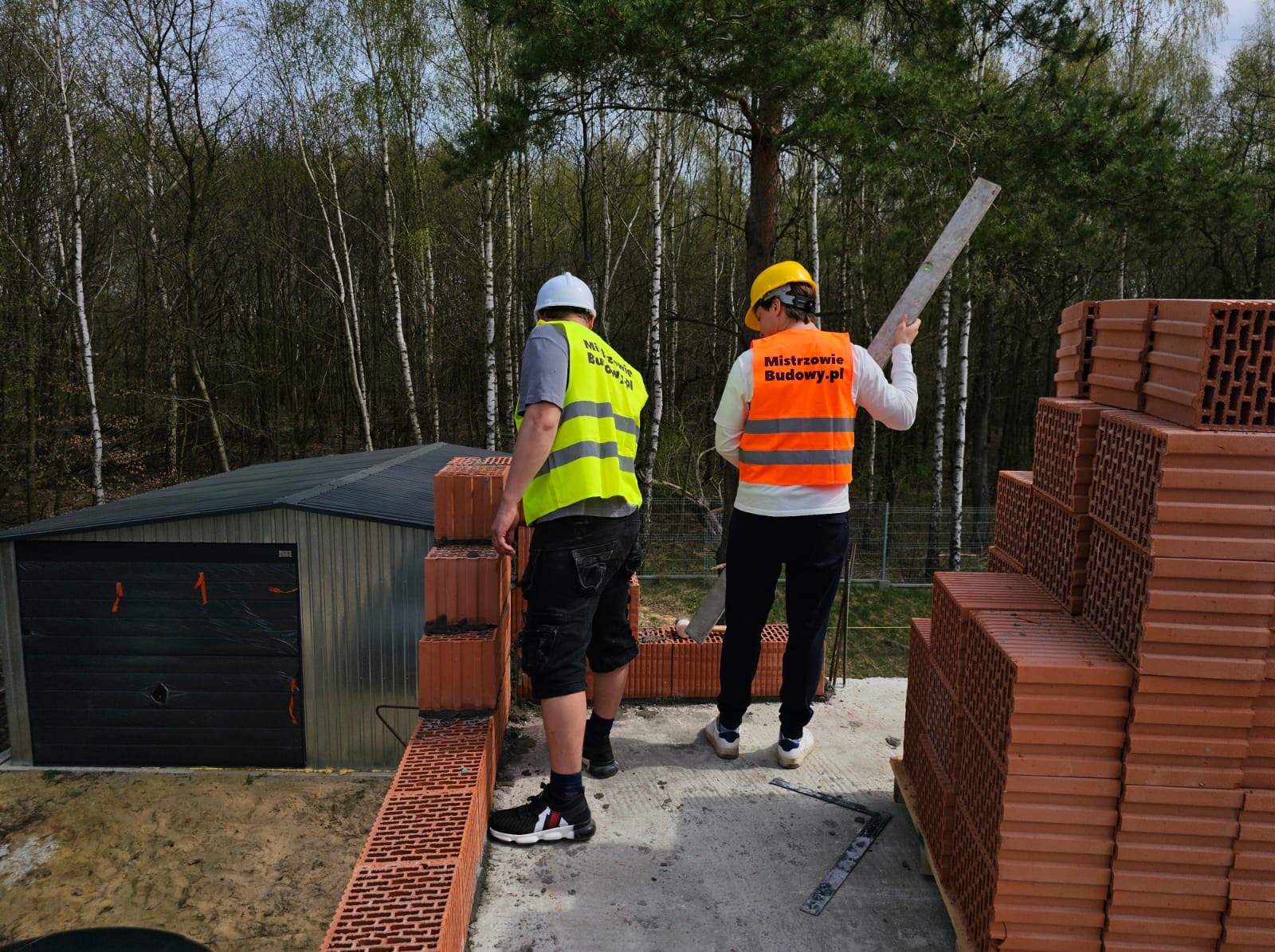 Dwóch pracowników budowlanych w odblaskowych kamizelkach, jeden z poziomicą, murują ścianę z czerwonej cegły na tle lasu i blaszanej konstrukcji garażu.