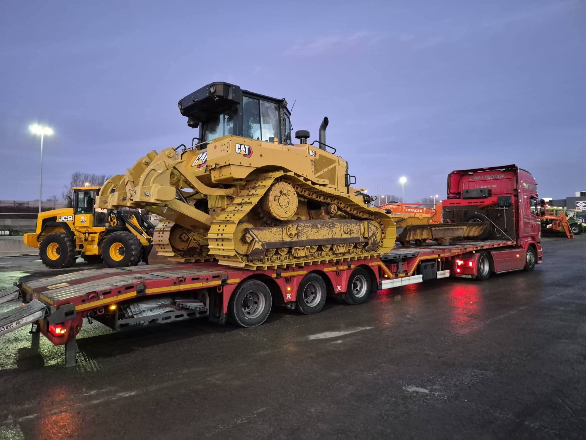 Transport niskopodwoziowy: czerwona ciężarówka Scania przewozi żółty spychacz CAT D6 na lawecie. W tle widoczny żółty JCB i pomarańczowy Hitachi.