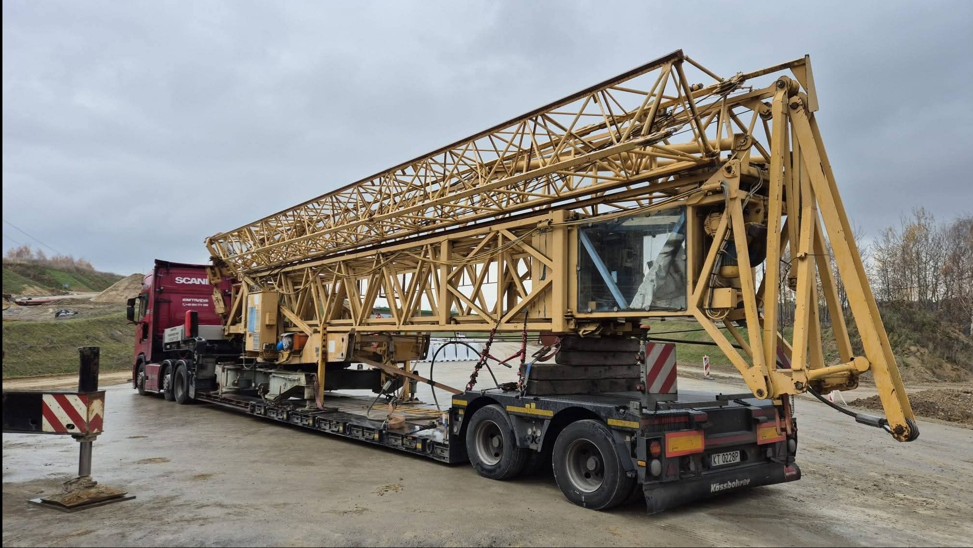 Żółty dźwig na niskopodwoziowej naczepie Scania, przygotowany do transportu. Widoczne zabezpieczenia ładunku i tablice ostrzegawcze. Pochmurne niebo w tle.