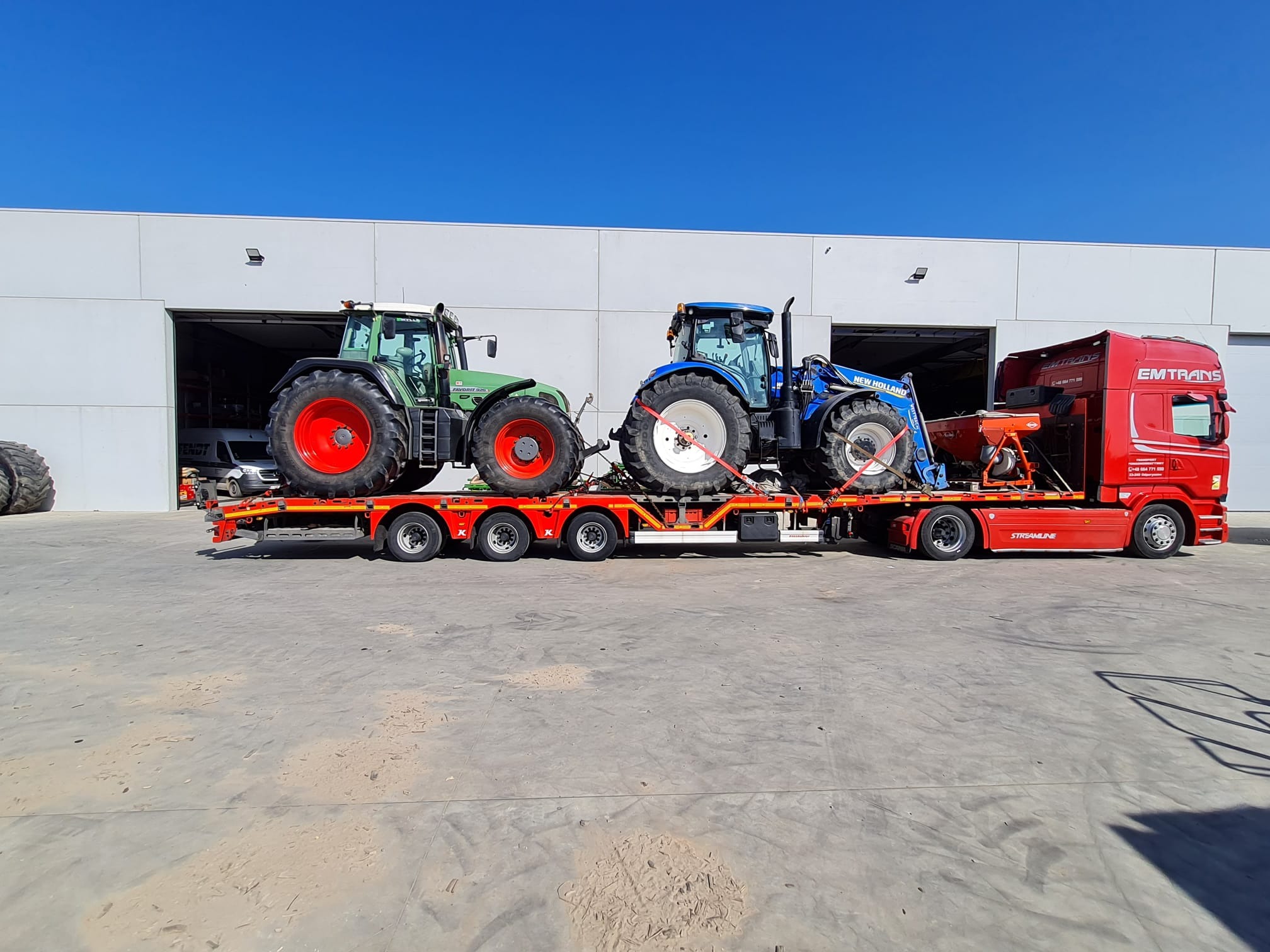 Czerwona ciężarówka EMTRANS przewozi dwa traktory (zielony i niebieski) na niskopodwoziowej platformie. Transport odbywa się na tle szarego budynku.