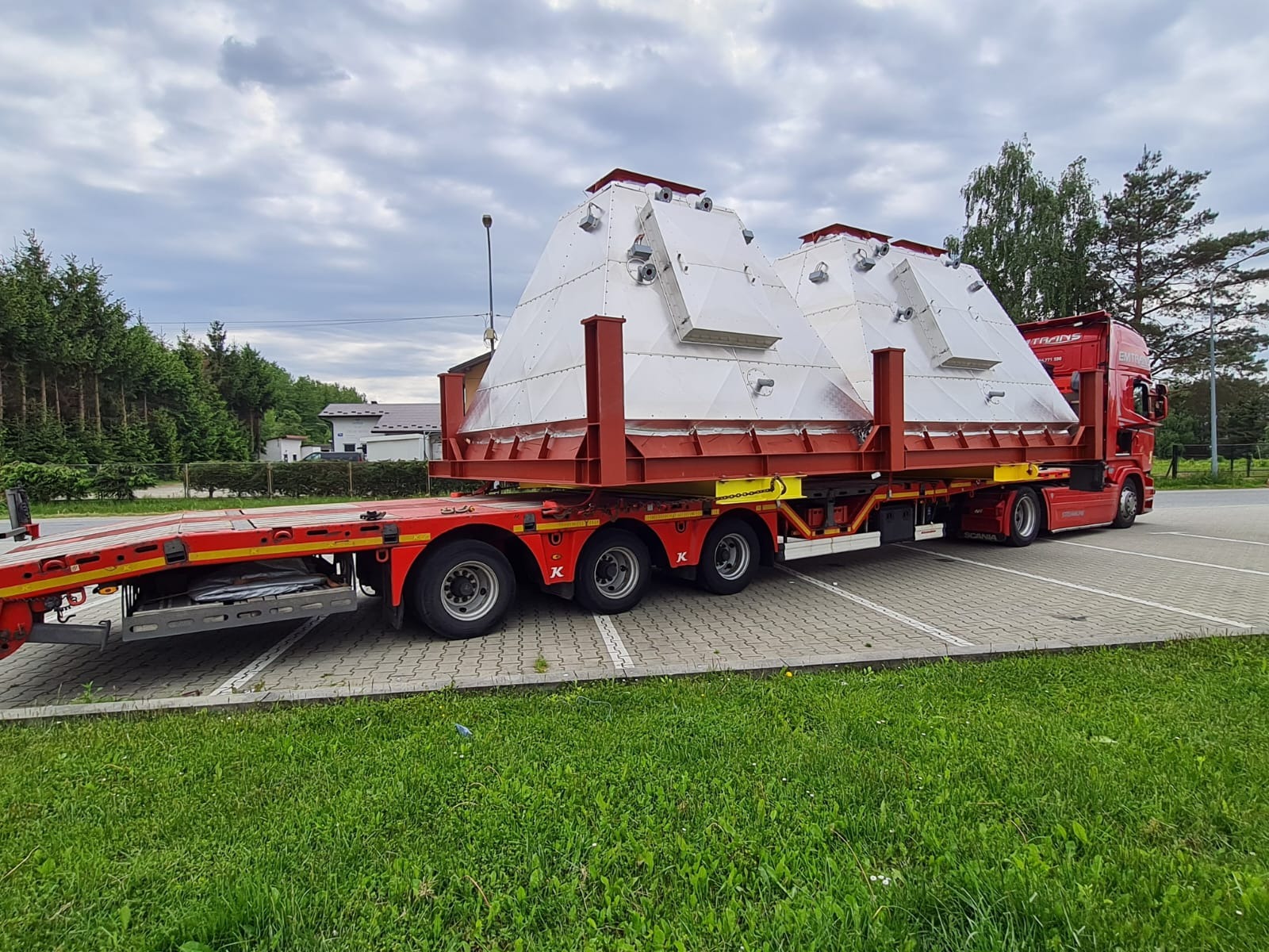 Czerwona ciężarówka z naczepą przewozi dwa duże, srebrne, piramidalne zbiorniki. Transport ponadgabarytowy na tle zieleni i pochmurnego nieba.