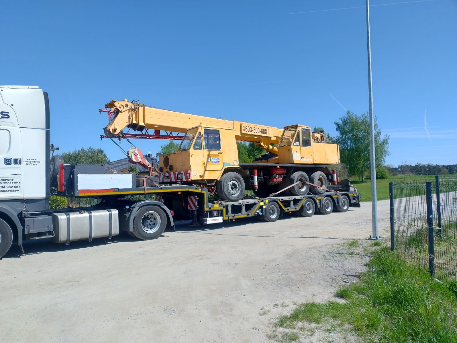 Żółty dźwig na niskopodwoziowej naczepie transportowany przez białą ciężarówkę. Błękitne niebo w tle. Widoczny numer telefonu na dźwigu.