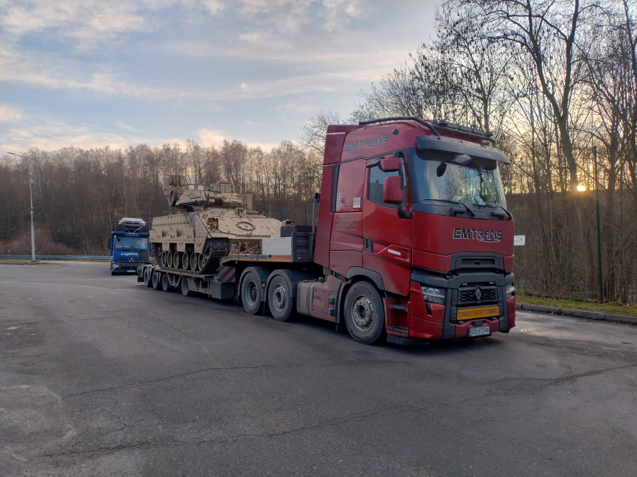 Czerwona ciężarówka EMTRANS przewozi na niskopodwoziowej platformie wojskowy pojazd opancerzony. W tle widoczny inny transportowiec i drzewa.
