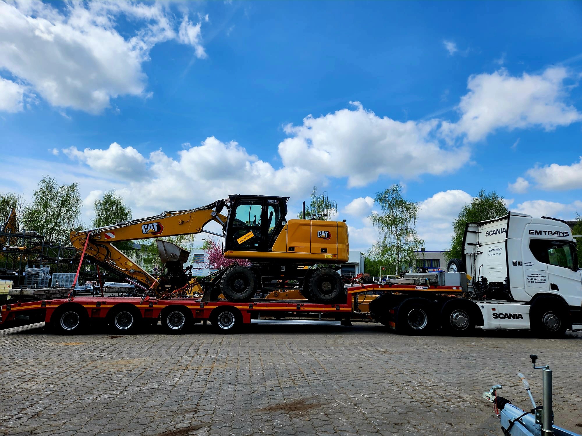 Żółta koparka CAT M316 na niskopodwoziowej naczepie, ciągniętej przez białą ciężarówkę Scania EMTRANS na tle błękitnego nieba z chmurami.