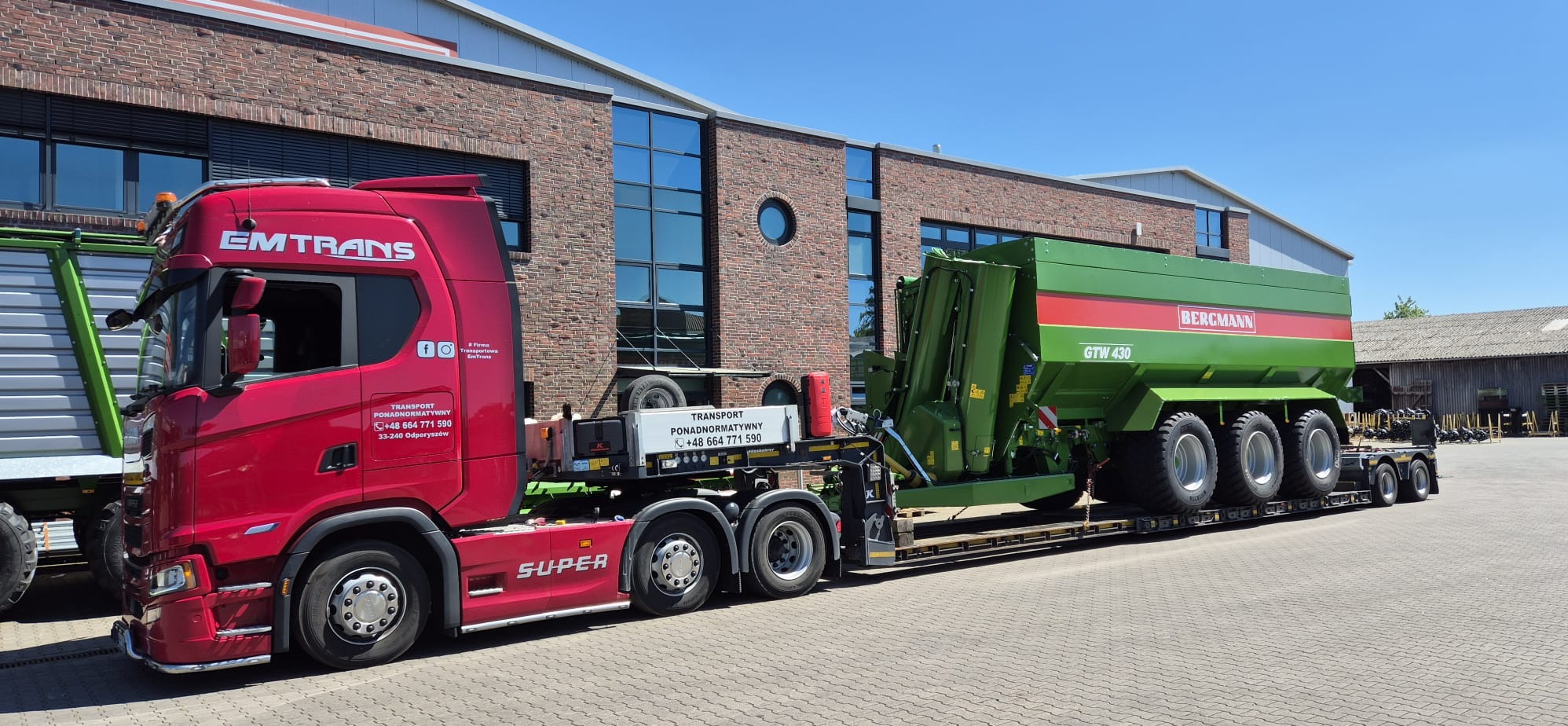 Czerwona ciężarówka EMTRANS przewozi na niskopodwoziowej platformie zielony, rolniczy wóz Bergmann GTW 430 na tle ceglanego budynku z dużymi oknami.
