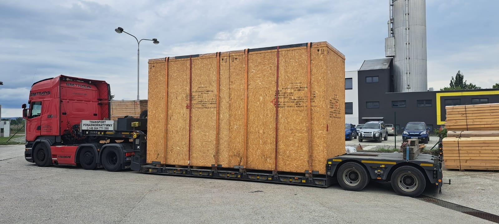 Czerwona ciężarówka z naczepą przewozi dużą, drewnianą skrzynię na tle przemysłowego krajobrazu z budynkiem i stertą drewna. Transport ponadgabarytowy.