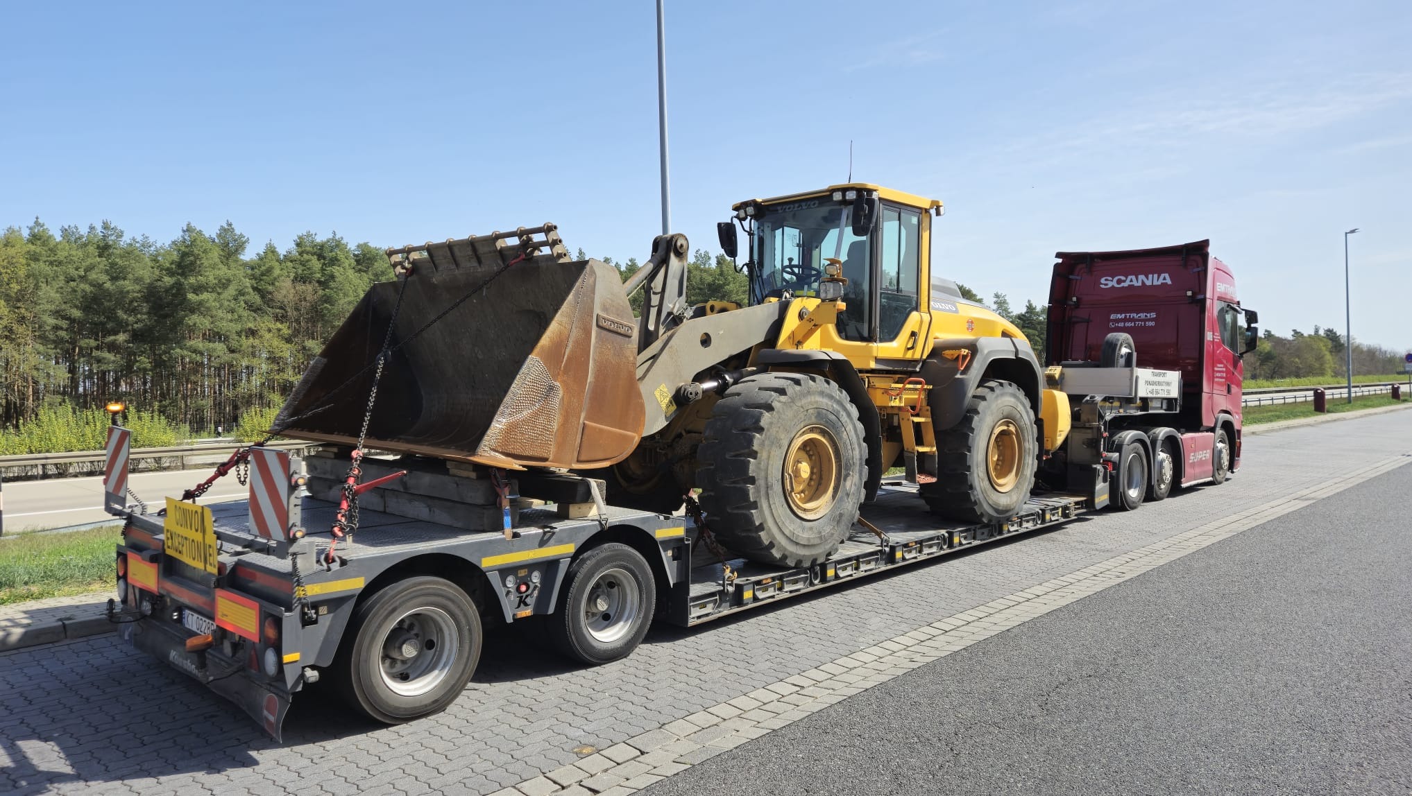 Transport niskopodwoziowy: żółta ładowarka Volvo na platformie, zabezpieczona łańcuchami, przewożona przez czerwoną ciężarówkę Scania. Transport ponadgabarytowy.