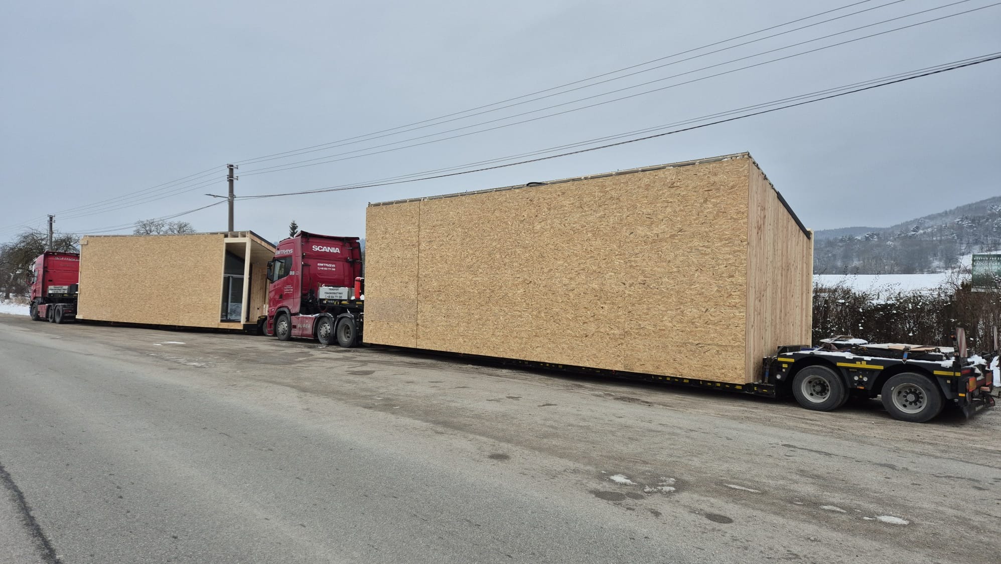 Czerwona ciężarówka Scania przewozi modułowy dom z płyty OSB na niskopodwoziowej naczepie. Widoczny drugi moduł i ciężarówka w tle, zimowy krajobraz.