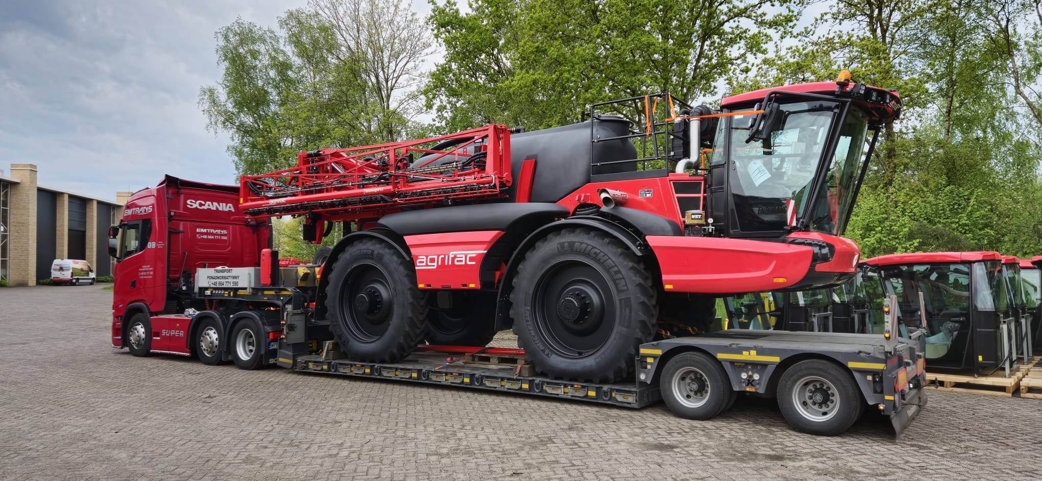 Czerwony ciągnik rolniczy Agrifac na niskopodwoziowej naczepie, transportowany przez czerwoną ciężarówkę Scania. Widoczne drzewa w tle.