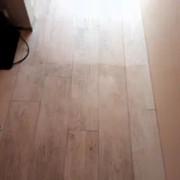 Jasne panele podłogowe, widoczne zabrudzenia i ślady po czyszczeniu, fragment czarnego odkurzacza w lewym dolnym rogu, ściana w tle.