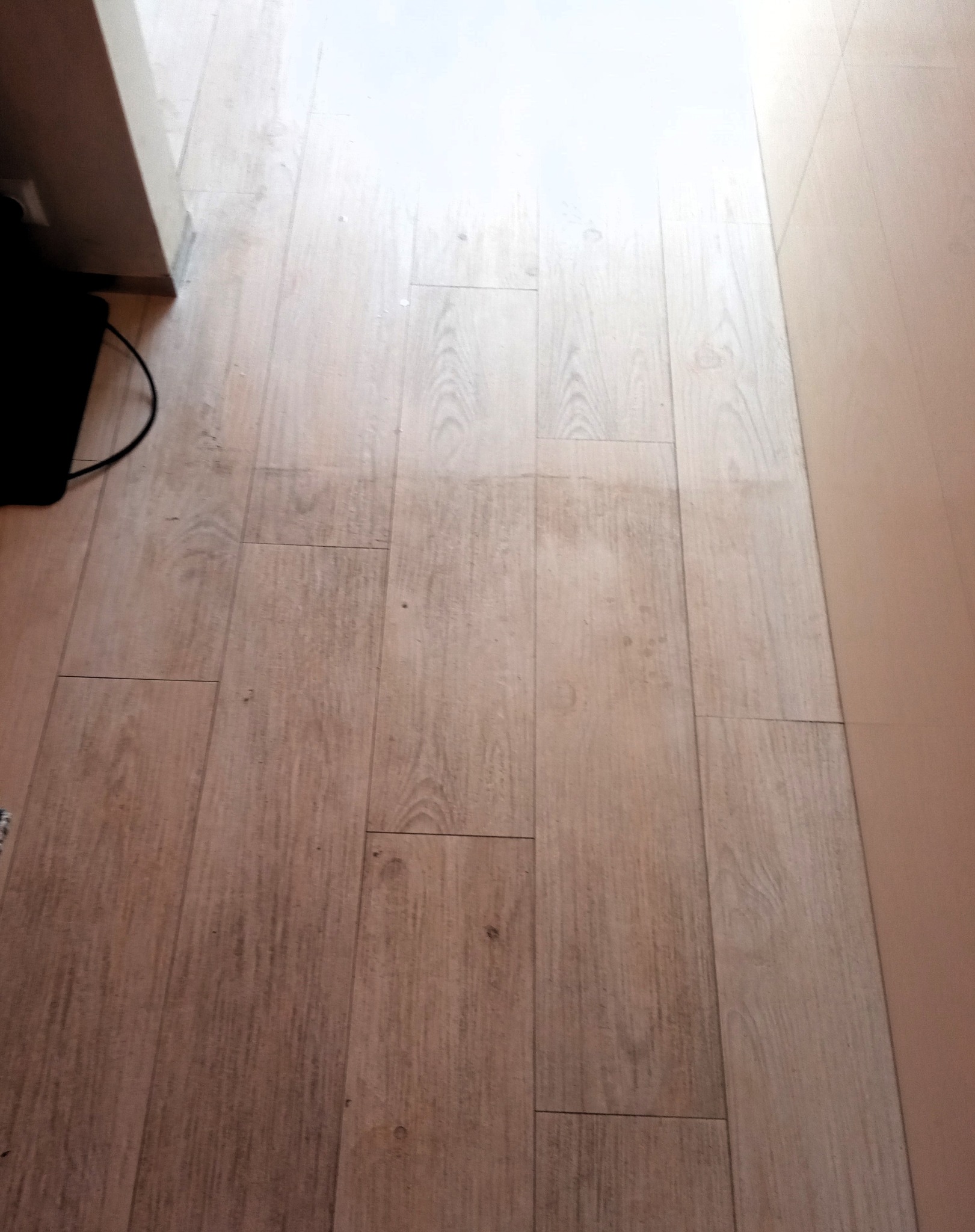 Jasne panele podłogowe, widoczne zabrudzenia i ślady po czyszczeniu, fragment czarnego odkurzacza w lewym dolnym rogu, ściana w tle.