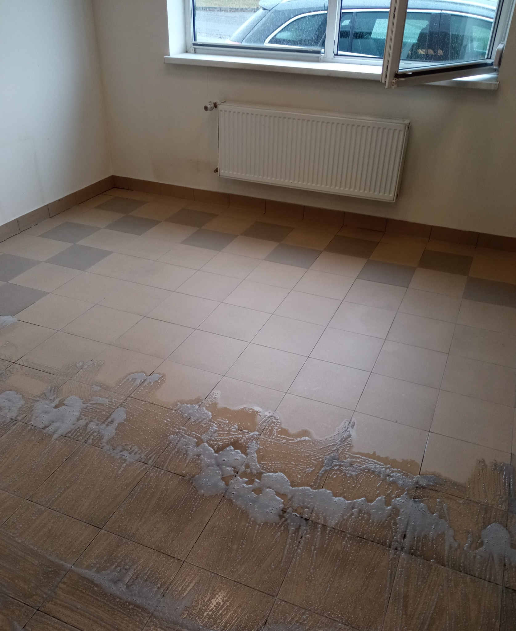 Płyn do czyszczenia na brudnej, kafelkowanej podłodze w pustym pokoju z grzejnikiem pod oknem. Widoczne ślady po remoncie.