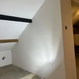 Poddasze po szpachlowaniu i malowaniu na biało, widoczne elementy konstrukcyjne dachu, surowa podłoga i lampa budowlana.