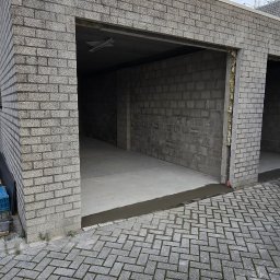 ŁUKASZ PIERSZCHAŁA GUM-BUD - Surowy, niewykończony garaż z szarej cegły, z widocznym ociepleniem między segmentami, betonowa podłoga i kostka brukowa przed wjazdem.