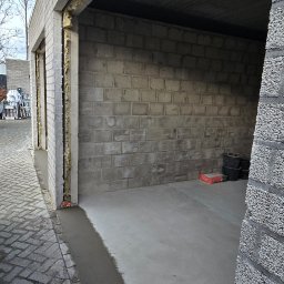 ŁUKASZ PIERSZCHAŁA GUM-BUD - Surowy garaż z bloczków betonowych, izolacją w szczelinach i świeżo wylaną wylewką, widok na zewnątrz z kostki brukowej.