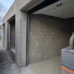 ŁUKASZ PIERSZCHAŁA GUM-BUD - Surowy garaż z szarej cegły bloczkowej, widoczne otwarte wnęki i częściowo zamontowana brama segmentowa, z elementami izolacji w otworach okiennych.