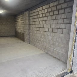 ŁUKASZ PIERSZCHAŁA GUM-BUD - Surowy garaż z betonowymi ścianami z pustaków, betonową podłogą i widoczną izolacją w otworze drzwiowym, oświetlony lampami jarzeniowymi na suficie.