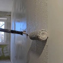 ŁUKASZ PIERSZCHAŁA GUM-BUD - Świeżo pomalowana ściana z widoczną fakturą farby, malowana wałkiem na teleskopowym kiju. Widoczne okno i fragment pomieszczenia w trakcie prac wykończeniowych.