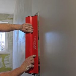 ŁUKASZ PIERSZCHAŁA GUM-BUD - Gładzenie ściany czerwoną szpachlą ParfaitLiss, widoczne dłonie z resztkami gładzi, fragment okna i surowej ściany w tle. Praca w trakcie wykańczania wnętrza.