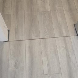 ŁUKASZ PIERSZCHAŁA GUM-BUD - Ułożone panele podłogowe w odcieniach szarości, imitujące drewno, widoczne progi drzwiowe. Układ paneli podłużny, tworzący jednolitą powierzchnię.