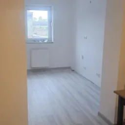 Jasne panele podłogowe imitujące drewno w nowo wykończonym pokoju z białymi ścianami. Widok z sąsiedniego pomieszczenia, okno z grzejnikiem w tle.