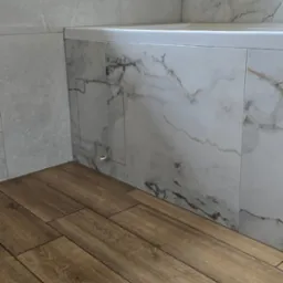 Wykończona łazienka: brązowe płytki imitujące drewno na podłodze, marmurowe płytki na ścianach i obudowie wanny. Widoczny fragment okna.