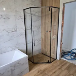 Wykończona łazienka z kabiną prysznicową typu walk-in z czarnymi profilami, drewnopodobną podłogą i marmurowymi płytkami ściennymi. Widoczna wanna i surowy otwór drzwiowy.