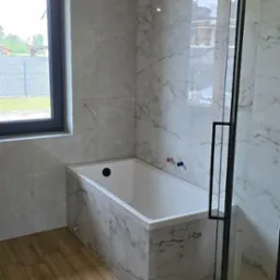 Nowoczesna łazienka z wanną obudowaną płytkami imitującymi marmur, drewnopodobna podłoga i czarna rama kabiny prysznicowej. Widok na okno.