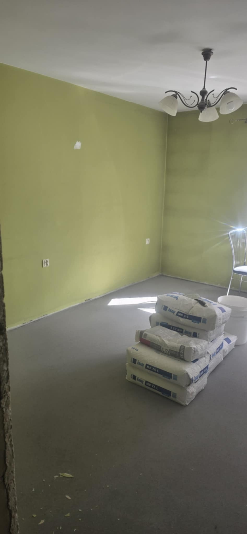 Wnętrze w trakcie prac: świeżo wylana wylewka podłogowa w pomieszczeniu z zielonymi ścianami. Widoczne worki z zaprawą i białe wiadro.