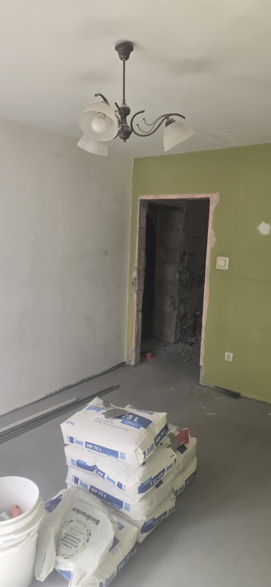 Pokój w trakcie prac: częściowo otynkowana ściana, materiały budowlane (worki z zaprawą Knauf MP 75 L), profil metalowy. Widoczne ślady ingerencji w ścianę.
