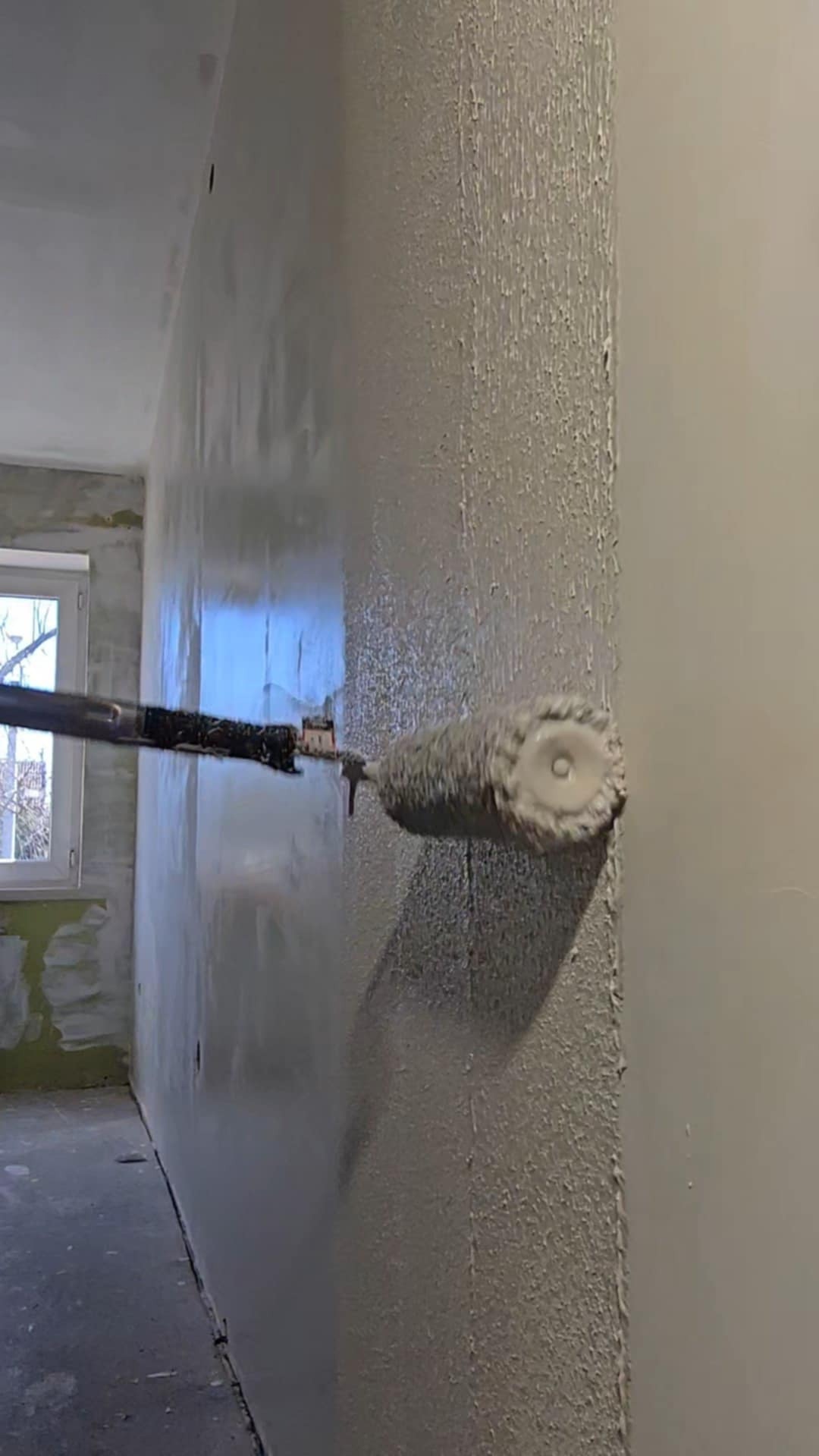 Świeżo pomalowana ściana z widoczną fakturą farby, malowana wałkiem na teleskopowym kiju. Widoczne okno i fragment pomieszczenia w trakcie prac wykończeniowych.