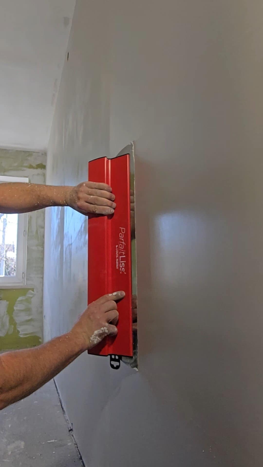 Gładzenie ściany czerwoną szpachlą ParfaitLiss, widoczne dłonie z resztkami gładzi, fragment okna i surowej ściany w tle. Praca w trakcie wykańczania wnętrza.