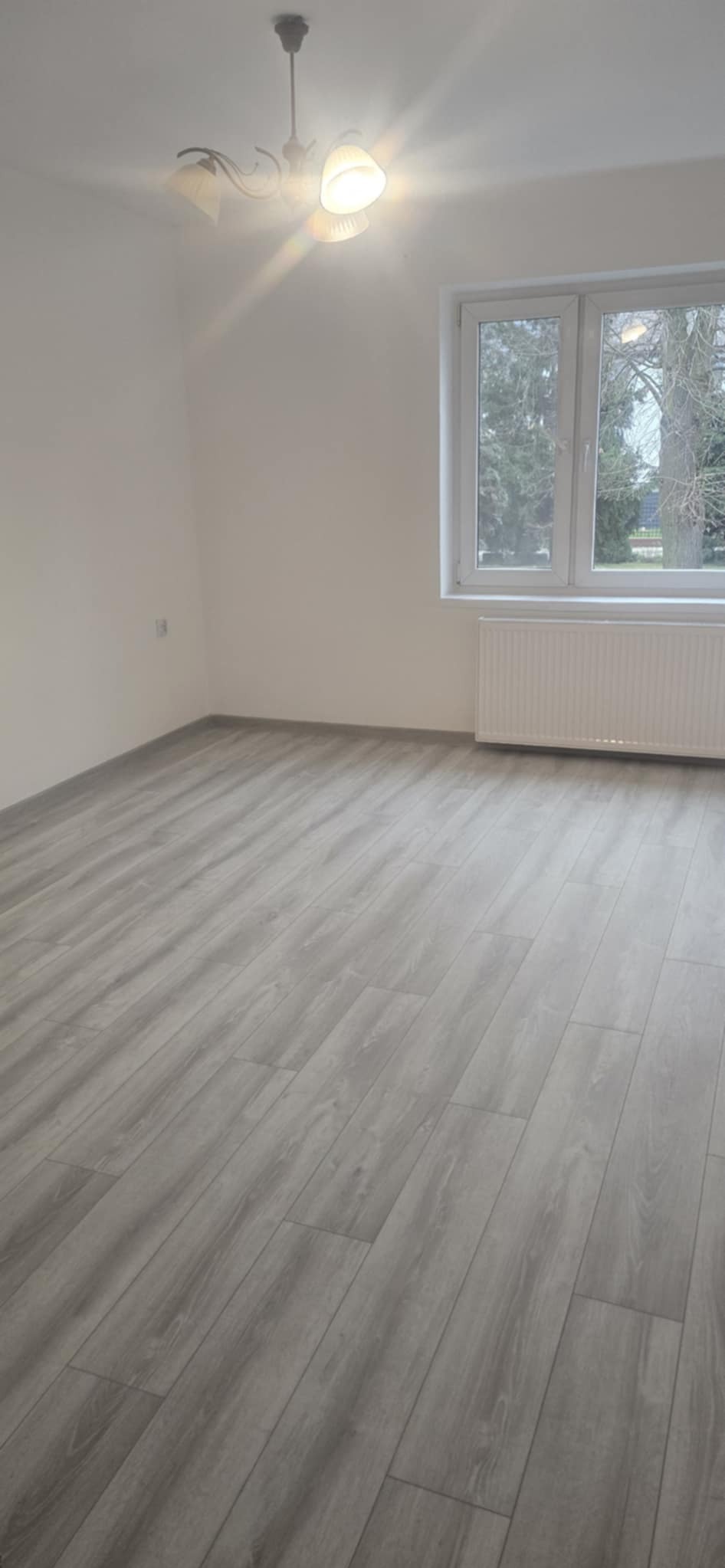 Jasne, puste wnętrze z panelami podłogowymi imitującymi drewno. Widoczne okno z grzejnikiem, lampa sufitowa i gładkie, białe ściany.