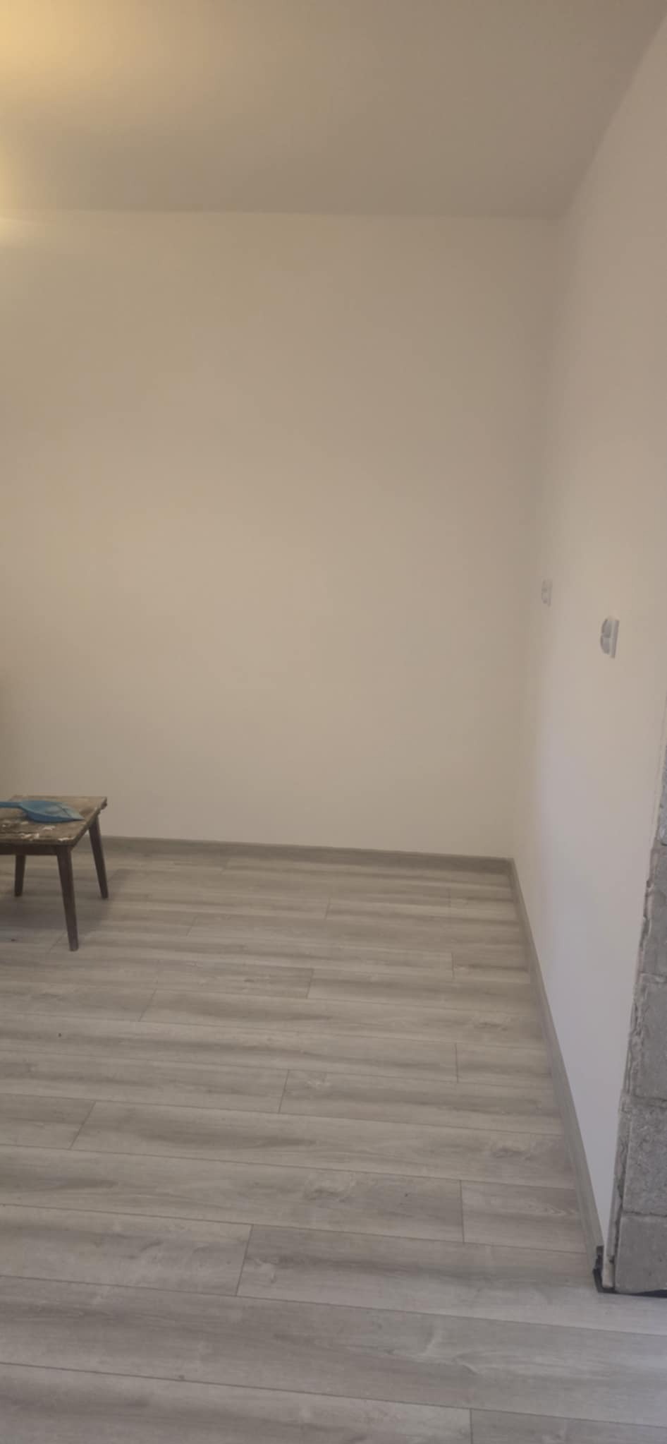 Jasne panele podłogowe w pokoju po remoncie, przy ścianie drewniany taboret z niebieską szmatką. Widoczne gniazdka elektryczne na ścianie.