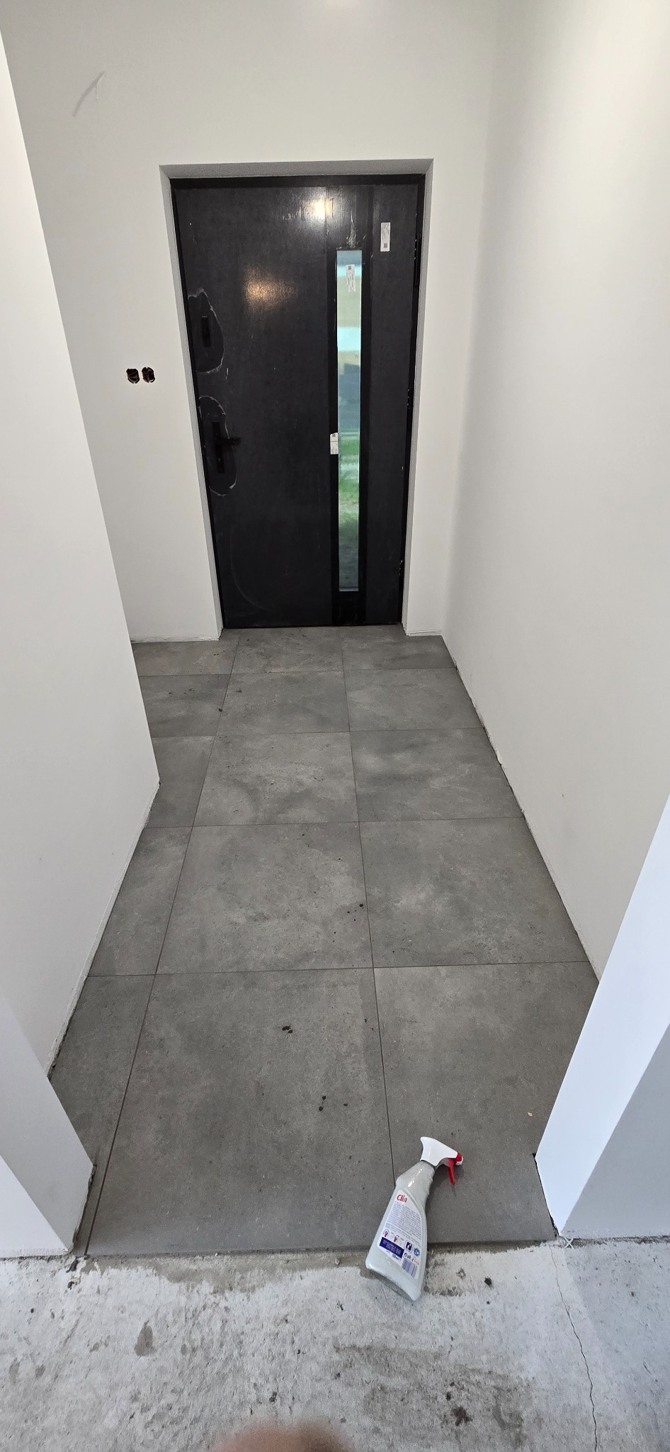 Wykończony korytarz z szarą podłogą z płytek. Widoczna czarna rama drzwi wejściowych. Na podłodze leży butelka ze środkiem czyszczącym.