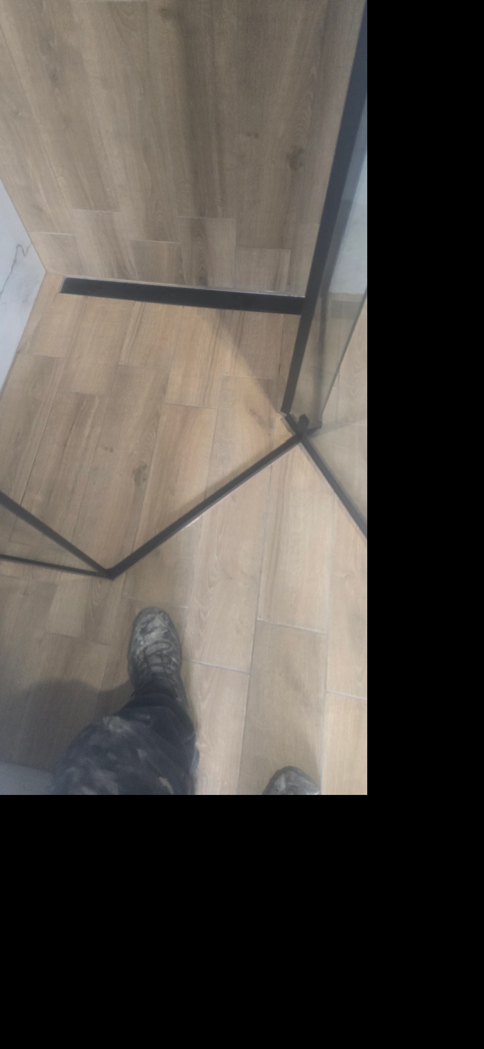 Bieżnikowana podłoga łazienki z odpływem liniowym, kabina prysznicowa z czarnymi profilami, fragment butów osoby fotografującej.