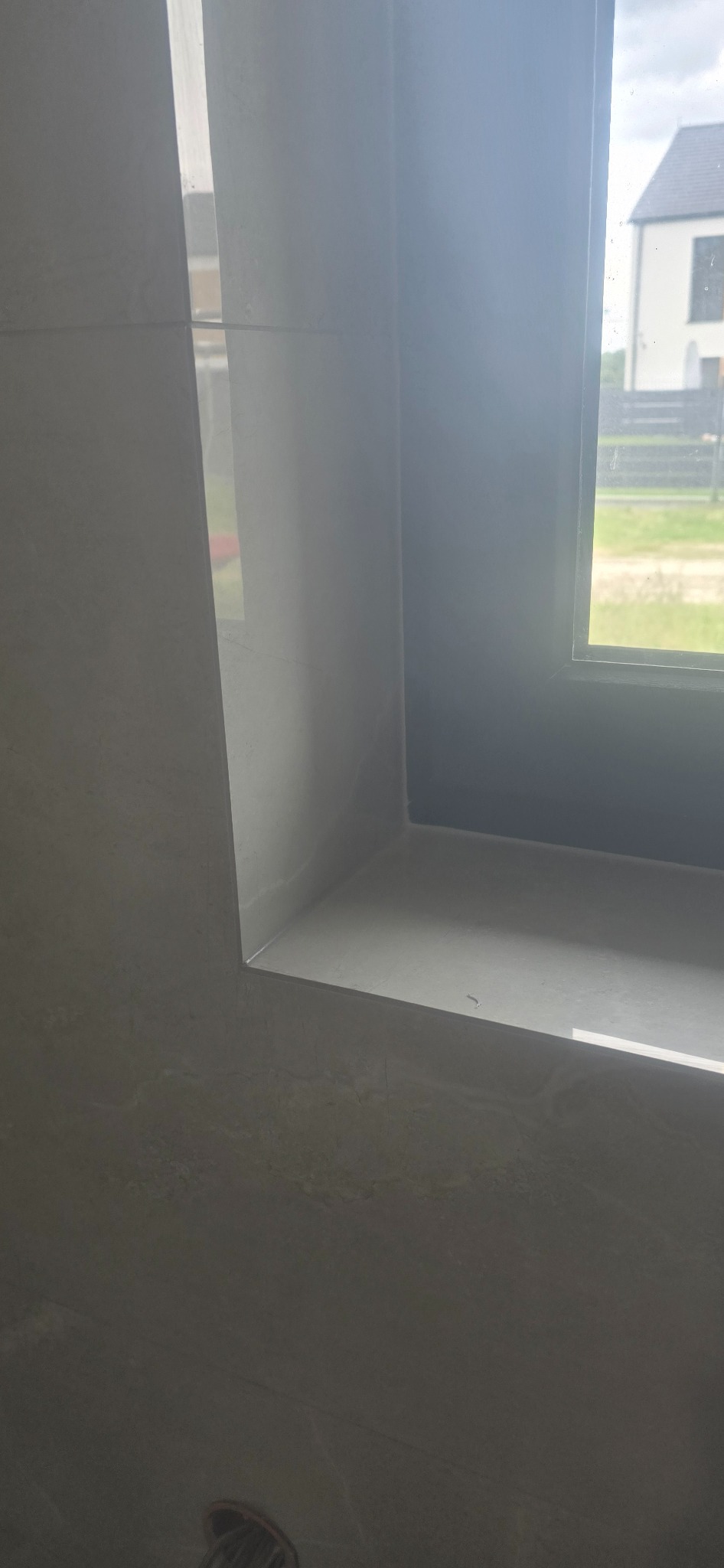 Wykończone wnęki okienne w łazience, obłożone jasnymi płytkami o delikatnej fakturze. Widok na zewnątrz przez okno. Detal wykończenia.