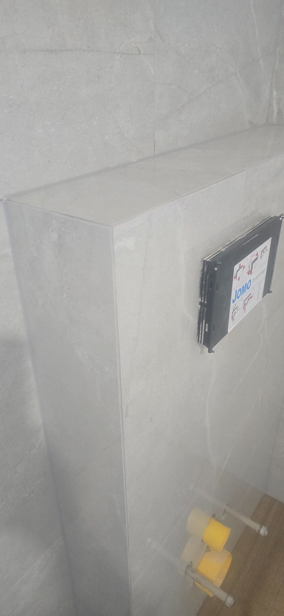 Wykończona obudowa stelaża WC z szarych płytek imitujących kamień. Widoczna instalacja podtynkowa JOMO i fragment podłogi w odcieniach brązu.