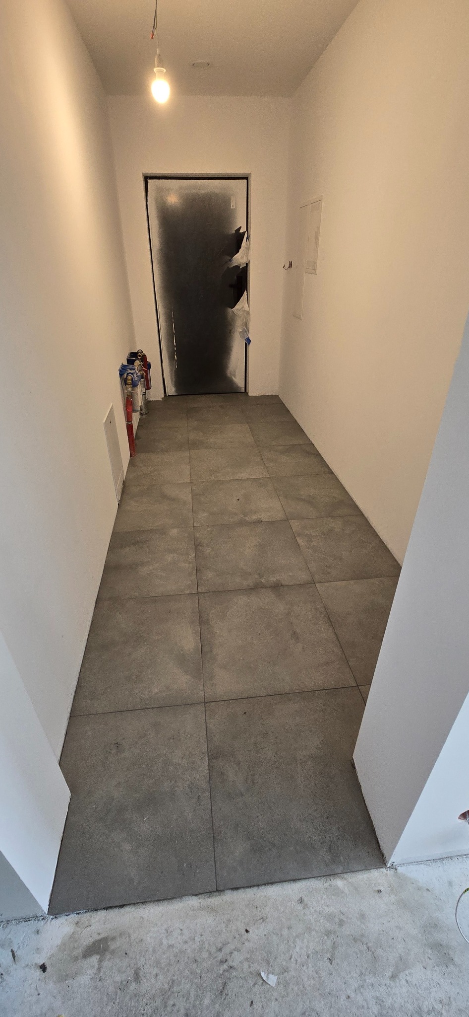 Szare, kwadratowe płytki podłogowe ułożone w korytarzu. Ściany białe, drzwi zabezpieczone folią. Widoczne rury i instalacje przy ścianie.